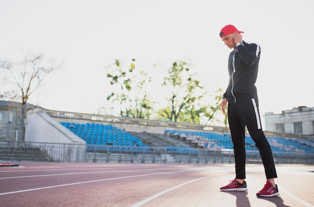 Apuesto joven atlético usando auriculares mirando hacia la pista en el estadio Atractivo corredor escuchando música durante el trote matutino y corriendo al aire libre Gente deportiva y estilo de vida