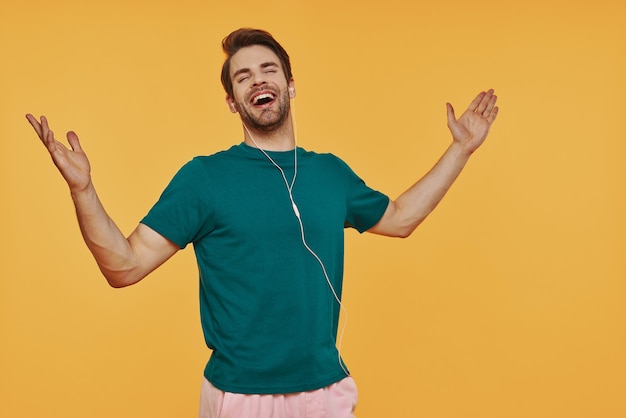 Apuesto joven en auriculares gesticulando y sonriendo mientras está de pie contra la pared amarilla