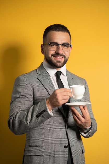 Apuesto joven barbudo en anteojos en ropa formal sosteniendo una taza de café aislada en la pared amarilla