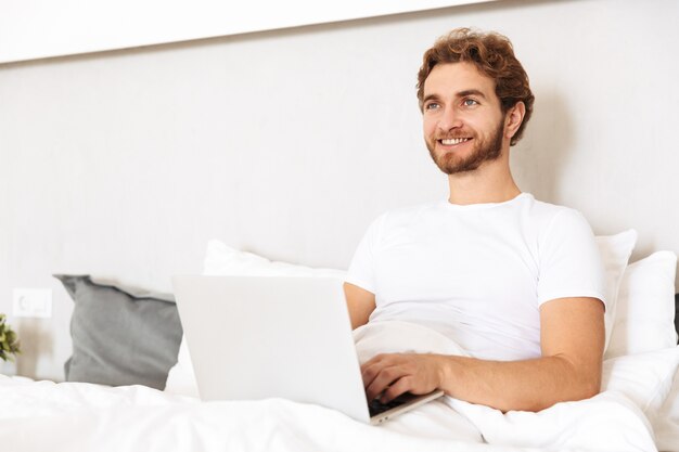 un apuesto joven barbudo en la cama en su casa usando la computadora portátil.