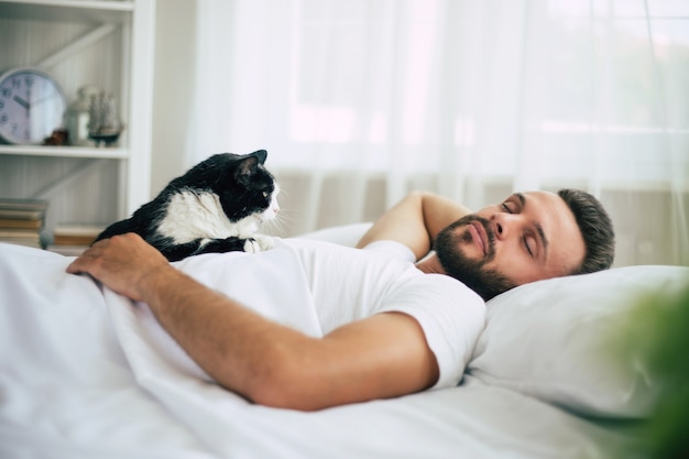 Apuesto joven barbudo está durmiendo en una cama grande blanca en un dormitorio cómodo