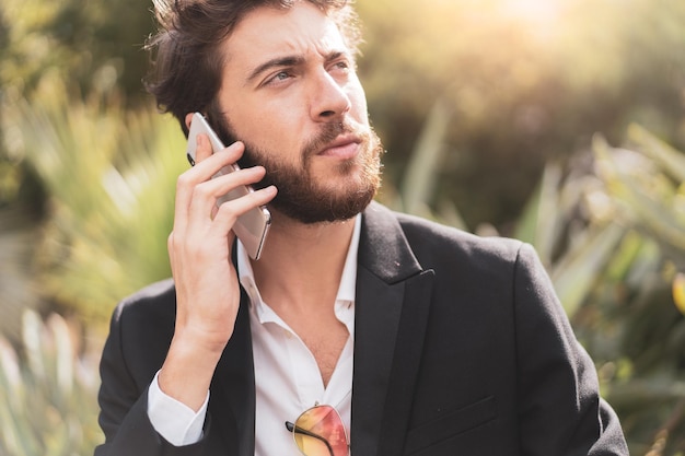 Apuesto joven barbudo en traje hablando por teléfono al aire libre.