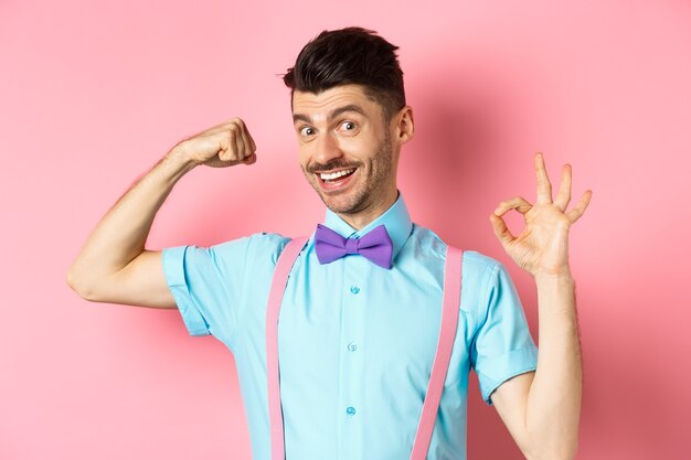 Apuesto joven con bigote y pajarita, flexión de bíceps, mostrando un brazo fuerte y signo bien, entrenamiento en el gimnasio, de pie sobre fondo rosa.