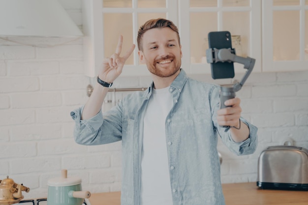 Apuesto joven brunete sonriente con rastrojo haciendo selfie en la cocina