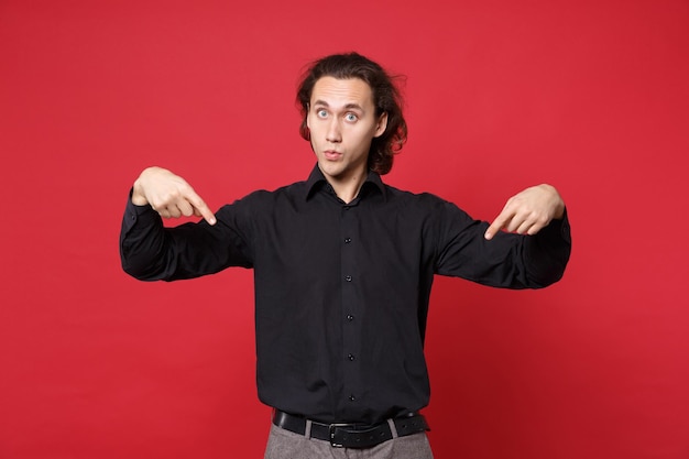 Un apuesto joven de cabello largo y rizado con camisa negra posando aislado en un retrato de estudio de fondo de pared roja. Concepto de estilo de vida de emociones sinceras de personas Mock up apuntando con el dedo en el espacio de copia
