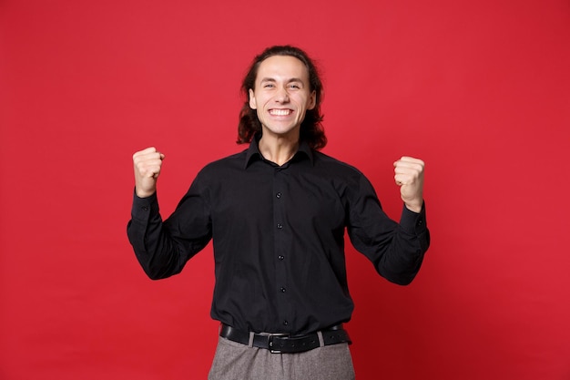 Un apuesto joven de cabello largo y rizado con camisa negra posando aislado en un retrato de estudio de fondo de pared roja. Gente emociones sinceras concepto de estilo de vida. Simulacros de espacio de copia. Haciendo gesto de ganador.