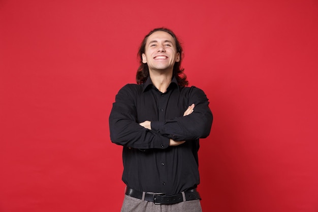 Un apuesto joven de cabello largo y rizado con camisa negra posando aislado en un retrato de estudio de fondo de pared roja. Gente emociones sinceras concepto de estilo de vida. Simulacros de espacio de copia. tomados de las manos cruzadas