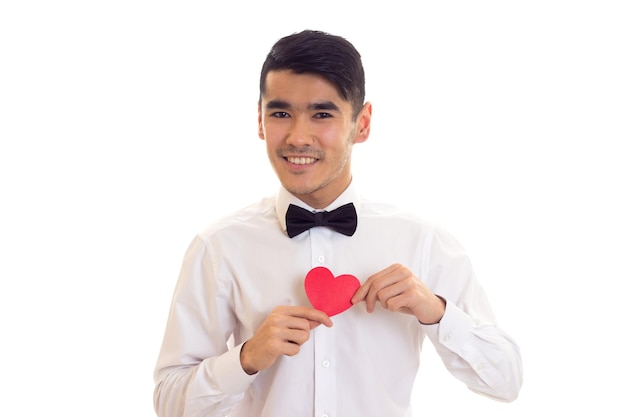 Apuesto joven con cabello negro en camiseta blanca con corbatín negro sosteniendo un corazón de papel rojo