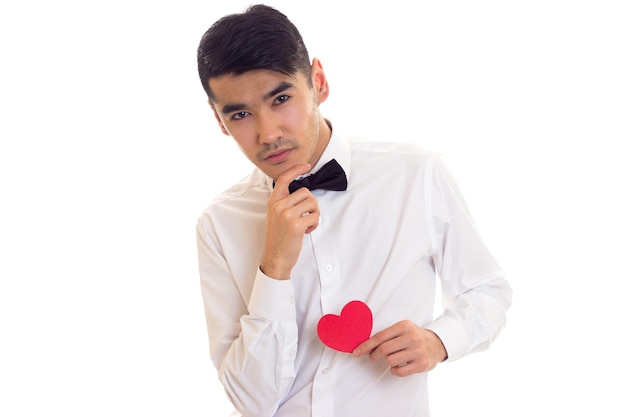 Apuesto joven con cabello oscuro en camiseta blanca con corbatín negro sosteniendo un corazón de papel rojo