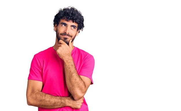 Apuesto joven con cabello rizado y oso con camiseta rosa casual con la mano en la barbilla pensando en la expresión pensativa de la pregunta sonriendo con el concepto de duda en la cara pensativa