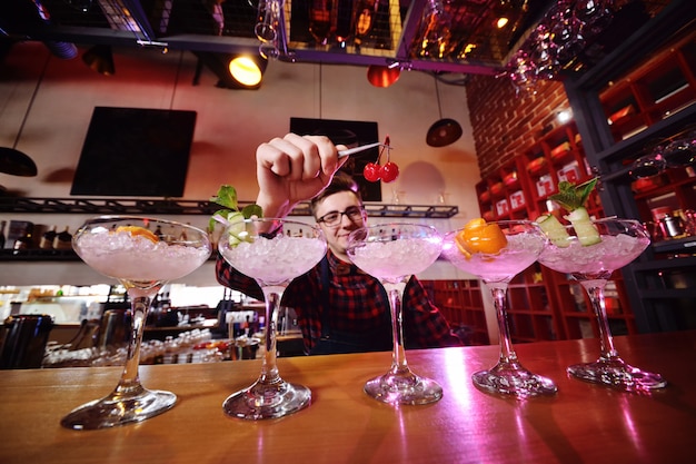 Un apuesto joven camarero prepara cócteles y pone cerezas para cócteles en la superficie de un bar o club nocturno.