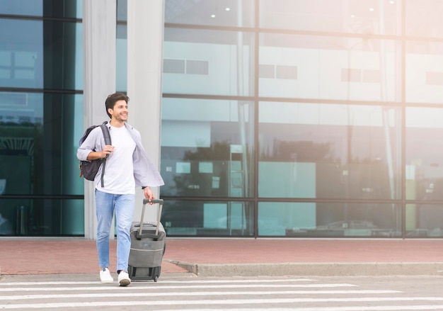 Apuesto joven caminando con equipaje fuera del aeropuerto con maleta