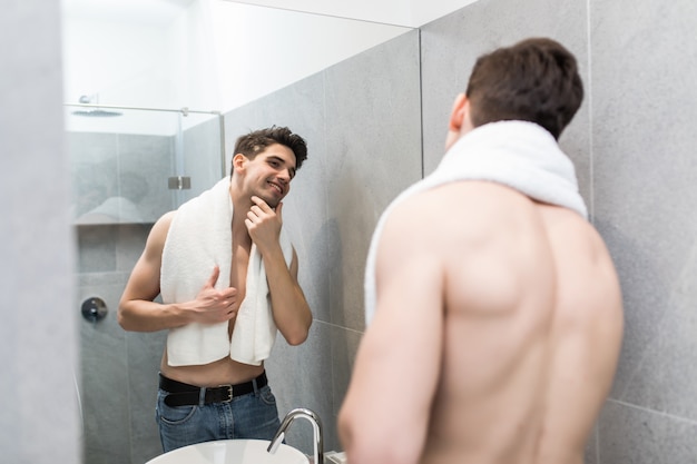 Apuesto joven sin camisa mirando en el espejo en el baño