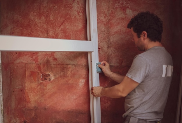 Un apuesto joven caucásico lava los marcos de las ventanas blancas con una esponja y jabón contra una pared roja