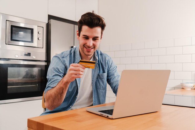 Apuesto joven caucásico sonriendo y sosteniendo una tarjeta de crédito para pagar una compra en línea usando una computadora portátil en la cocina de casa Concepto de finanzas bancarias