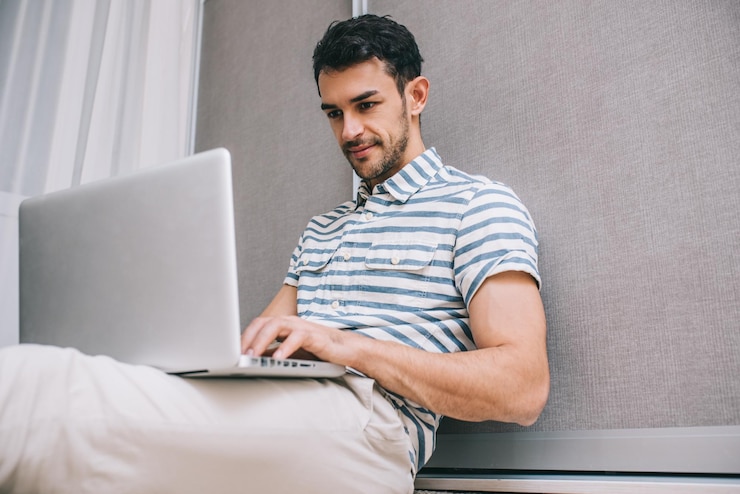 Foto apuesto joven caucásico sonriente con camisa elegante sentado en el suelo en casa con un ordenador portátil en su regazo aviso en papel para el inicio de negocios concepto de personas y estilo de vida