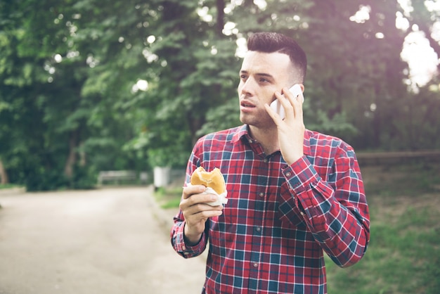 Apuesto joven comiendo bocadillo autdoor. El esta sosteniendo un telefono