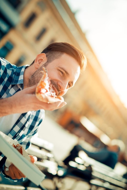 Apuesto joven comiendo una rebanada de pizza afuera en la calle.