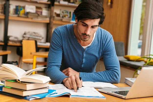 Foto apuesto joven confiado sentado en el escritorio de la biblioteca, trabajando / estudiando, usando la computadora portátil