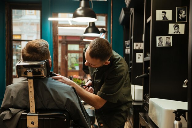 apuesto joven conseguir corte de pelo de barba por peluquero mientras está sentado en una silla en la barbería.