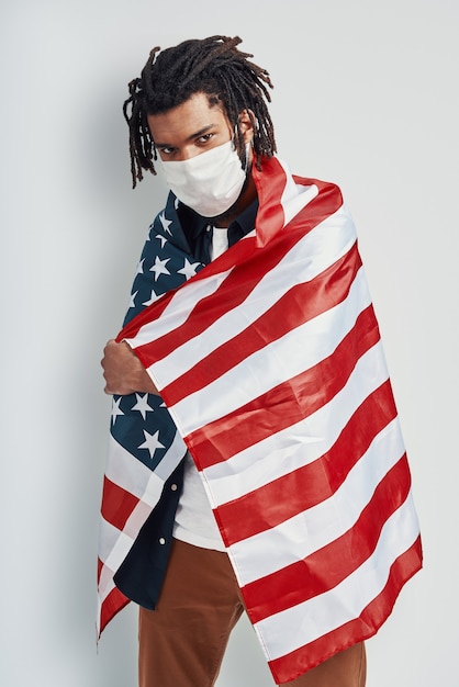 Apuesto joven cubierto con la bandera estadounidense con mascarilla médica y mirando a la cámara mientras está de pie contra la pared gris
