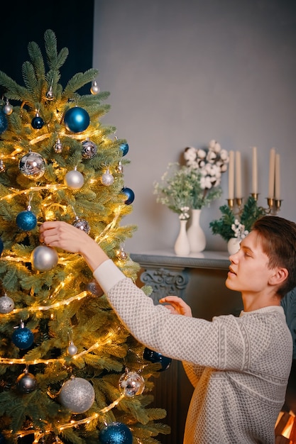 Apuesto joven cuelga la bola de plata en el árbol de navidad decoraciones coloridas y guirnaldas brillantes en n ...