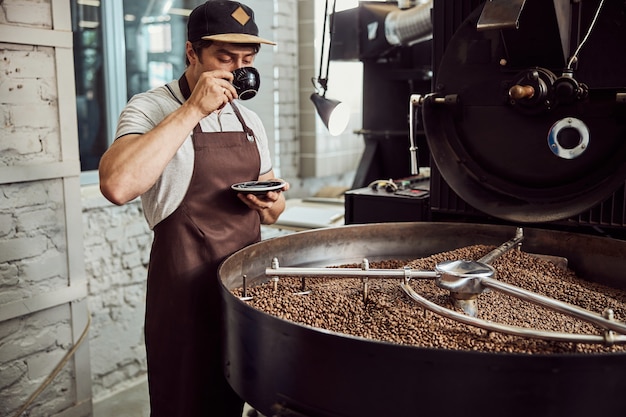 Apuesto joven en delantal degustación de bebidas calientes mientras está de pie junto a un equipo profesional de tostado de café