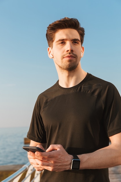 Apuesto joven deportista escuchando música con auriculares mediante teléfono móvil.