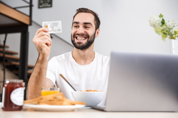 Apuesto joven desayunando mientras está sentado en la cocina, comprando con computadora portátil y tarjeta de crédito