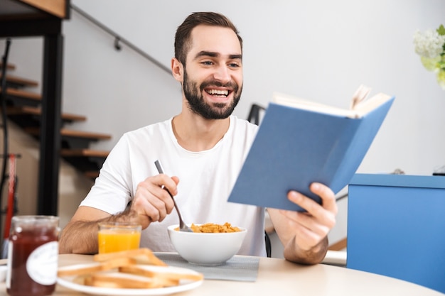 Apuesto joven desayunando mientras está sentado en la cocina, leyendo un libro