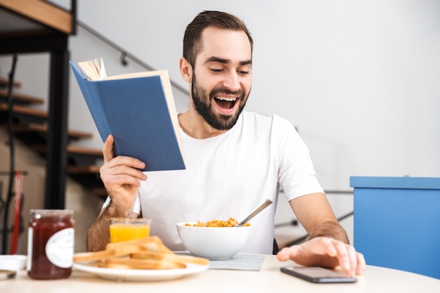 Apuesto joven desayunando mientras está sentado en la cocina, leyendo un libro