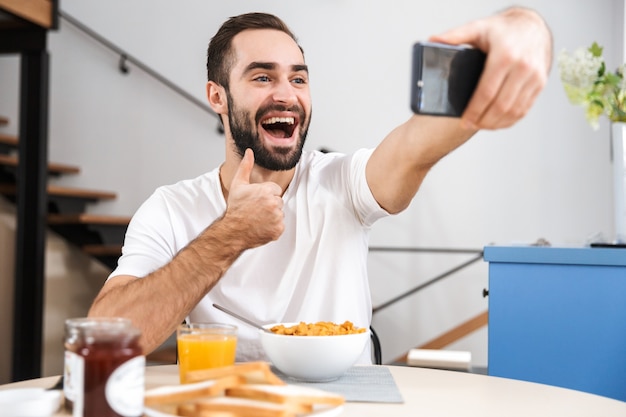 Apuesto joven desayunando mientras está sentado en la cocina, tomando un selfie