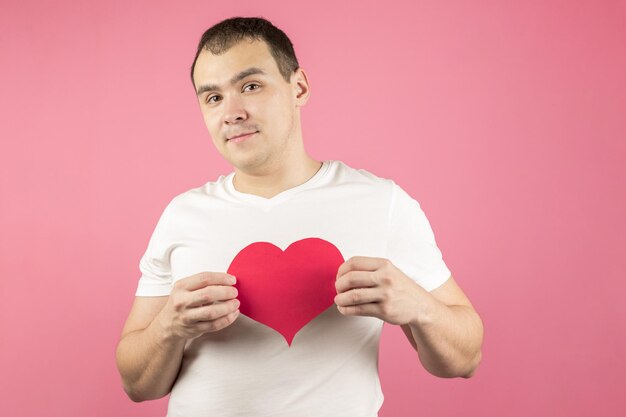 Un apuesto joven con una dulce sonrisa en una camiseta blanca sostiene un gran corazón de papel rojo o un fondo rosa de San Valentín Concepto del día de San Valentín declaración de amor amor felicidad