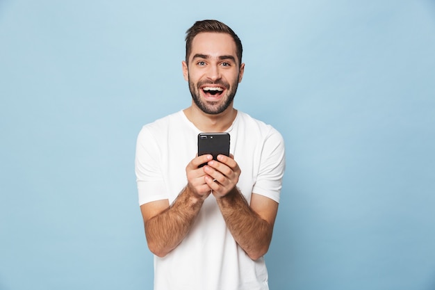 apuesto joven emocionado mediante teléfono móvil aislado sobre la pared azul.