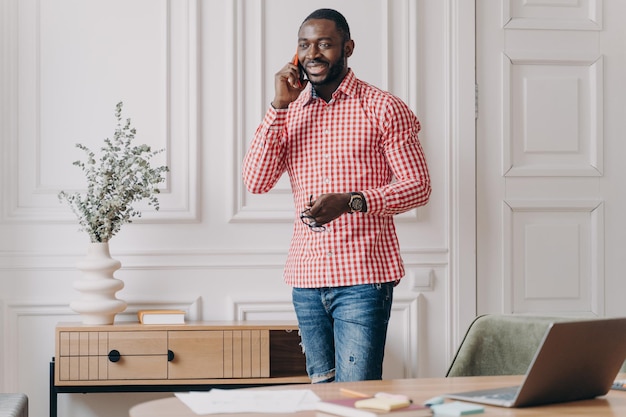 Apuesto joven empresario africano exitoso en ropa casual disfrutando de una llamada telefónica usando un teléfono inteligente