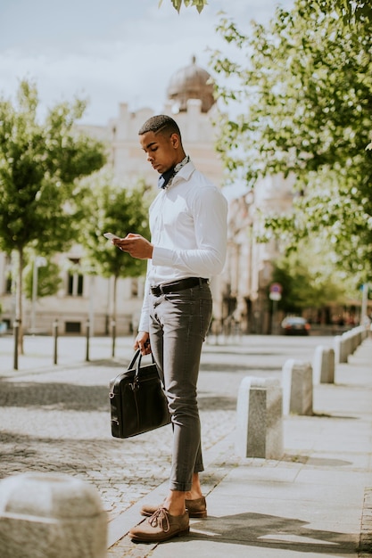 Apuesto joven empresario afroamericano mediante un teléfono móvil mientras espera un taxi en la calle