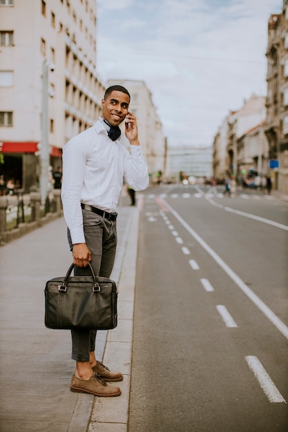 Apuesto joven empresario afroamericano mediante un teléfono móvil mientras espera un taxi esperando un taxi en una calle