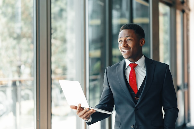 Apuesto joven empresario afroamericano en traje clásico sosteniendo una computadora portátil y sonriendo