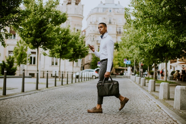 Apuesto joven empresario afroamericano usando un teléfono móvil mientras ceossing una calle