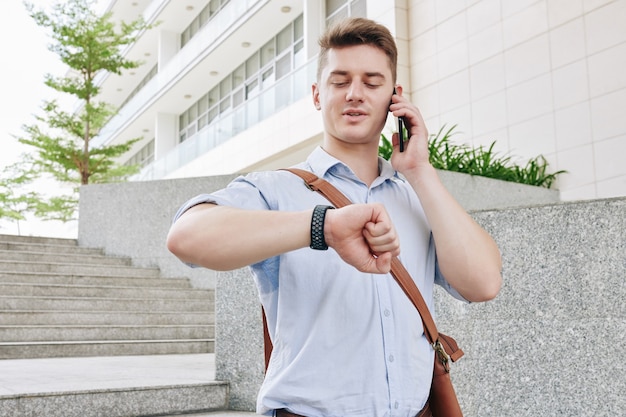 Apuesto joven empresario caminando por la calle, hablando por teléfono y controlando el tiempo en su reloj de pulsera