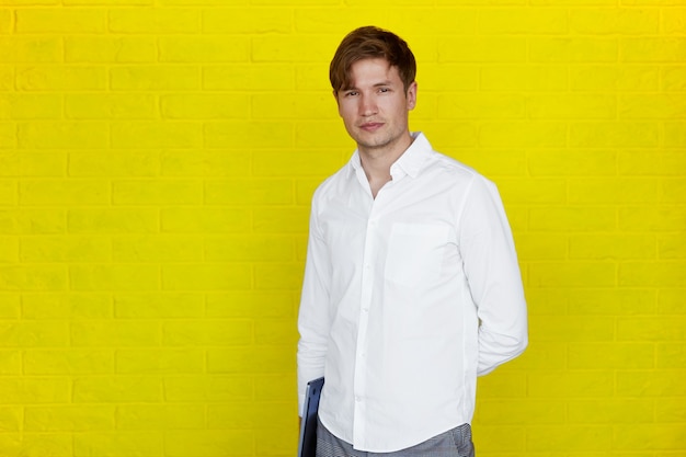 Apuesto joven empresario en camisa sosteniendo un portátil, mirando a cámara, sobre fondo amarillo.