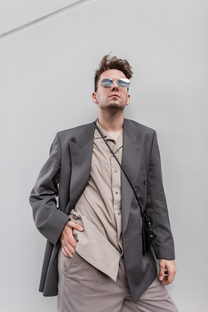 Apuesto joven empresario con cerdas y gafas en ropa de moda con camisa y chaqueta se encuentra cerca de fondo gris. Moda y estilo masculino casual