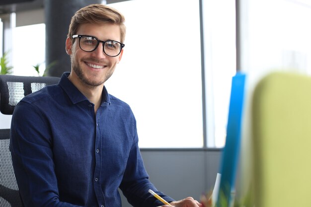 Apuesto joven empresario mirando a cámara y sonriendo mientras está sentado en la oficina.