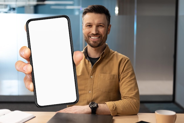 Apuesto joven empresario mostrando smartphone en blanco mientras está sentado en el escritorio en la oficina