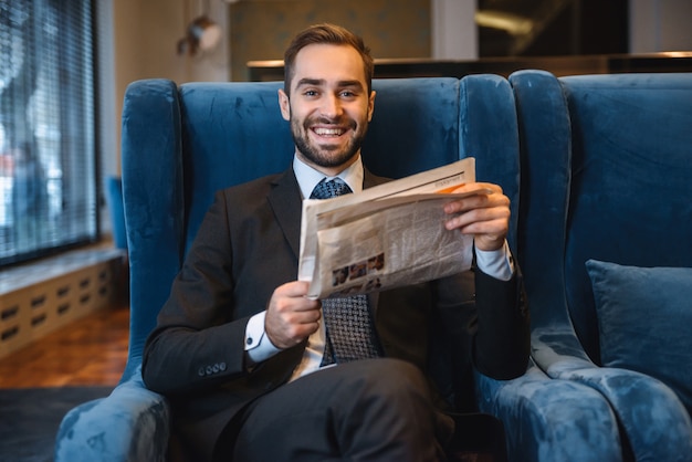 Apuesto joven empresario pensativo vistiendo traje sentado en el vestíbulo del hotel, leyendo el periódico