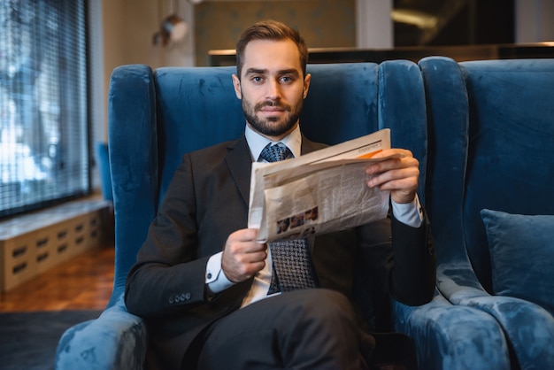 Apuesto joven empresario pensativo vistiendo traje sentado en el vestíbulo del hotel, leyendo el periódico