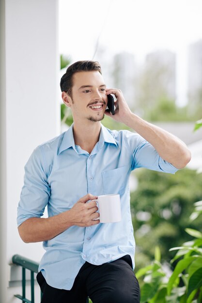 Apuesto joven empresario de pie en el balcón de la oficina, tomando café por la mañana y hablando por teléfono con su colega