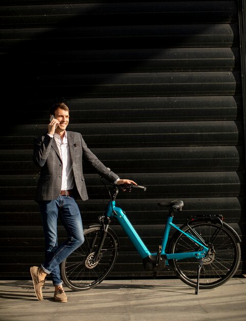 Apuesto joven empresario de pie junto a la bicicleta eléctrica y con móvil