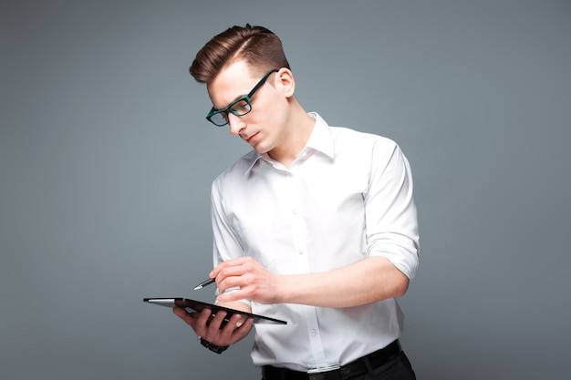 Apuesto joven empresario en reloj costoso, gafas negras y camisa blanca sostenga tableta y bolígrafo