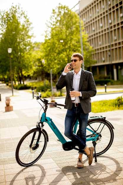 Apuesto joven empresario mediante teléfono móvil por la bicicleta eléctrica con taza de café para llevar